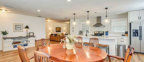 A modern kitchen with white cabinets, stainless steel appliances, and wooden chairs around a central dining table.