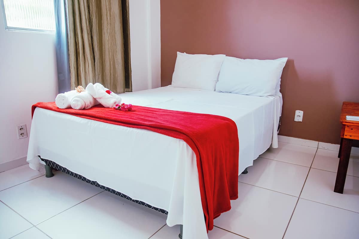 Un letto rifatto con lenzuola bianche e una coperta rossa, un comodino in legno e una finestra con tende.