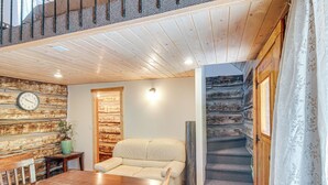 A cozy room with a wooden ceiling, a stone wall, a sofa, a dining table, and a staircase. - Bryce Canyon Basecamp: Historic Homesteader Cabin (Panguitch)