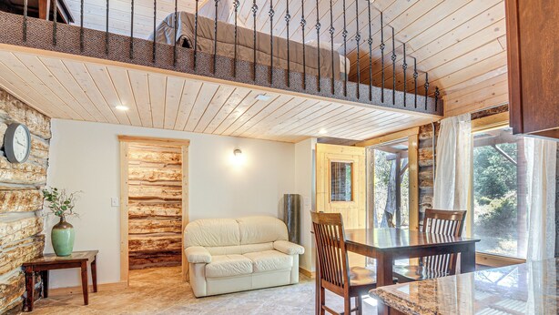 A cozy interior with a wooden ceiling, a stone wall, a beige sofa, a wooden dining table with chairs, and a large window offering a view of the outdoors. - Bryce Canyon Basecamp: Historic Homesteader Cabin (Panguitch)