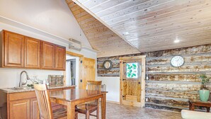 A kitchen with wooden cabinets, a dining table, and chairs. There is a clock on the wall and a door with a wooden frame. - Bryce Canyon Basecamp: Historic Homesteader Cabin (Panguitch)