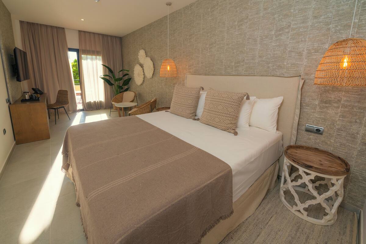 A spacious bedroom with a large bed, a bedside table, a chair, a small table, and a potted plant.