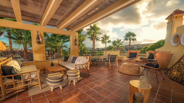 A sunlit patio with wicker furniture, a wooden table, and a view of palm trees and a distant town. - ALCAIDESA BOUTIQUE HOTEL (La Línea de la Concepción)