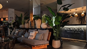 A modern lobby with a sofa, large potted plants, and a reception desk labeled "ALCAIDESA Boutique Hotel". - ALCAIDESA BOUTIQUE HOTEL (La Línea de la Concepción)