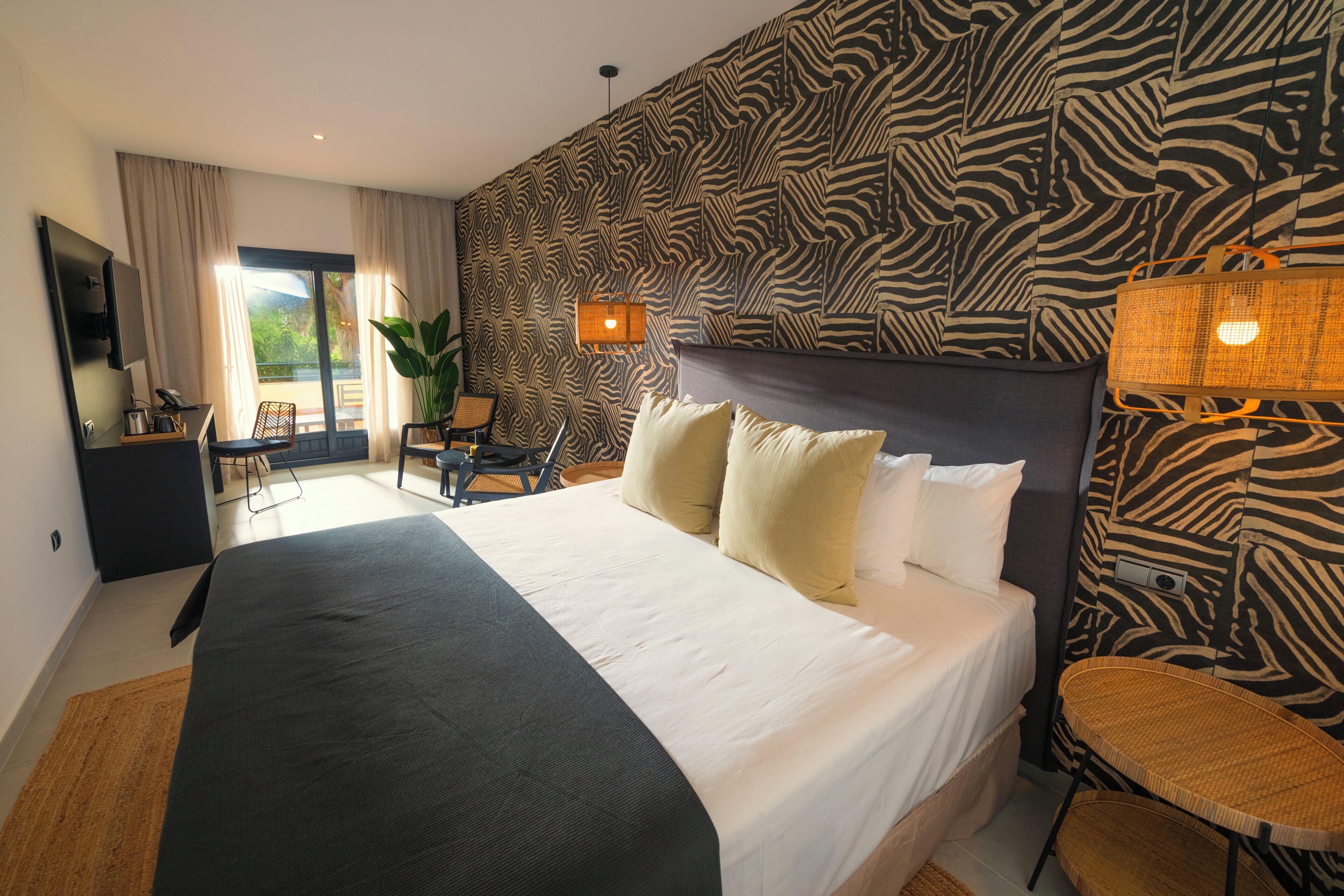 A hotel room with a large bed, a desk, a chair, a potted plant, and a zebra-patterned wall.