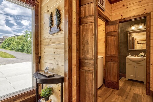 Ein Holzinterieur mit einem Fenster, einem Willkommensschild, einem kleinen Tisch mit einer Pflanze und einem Badezimmer mit Waschbecken und Spiegel.