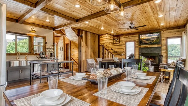 A spacious dining area with a long wooden table set for a meal, a kitchen with modern appliances, and a fireplace in the background.