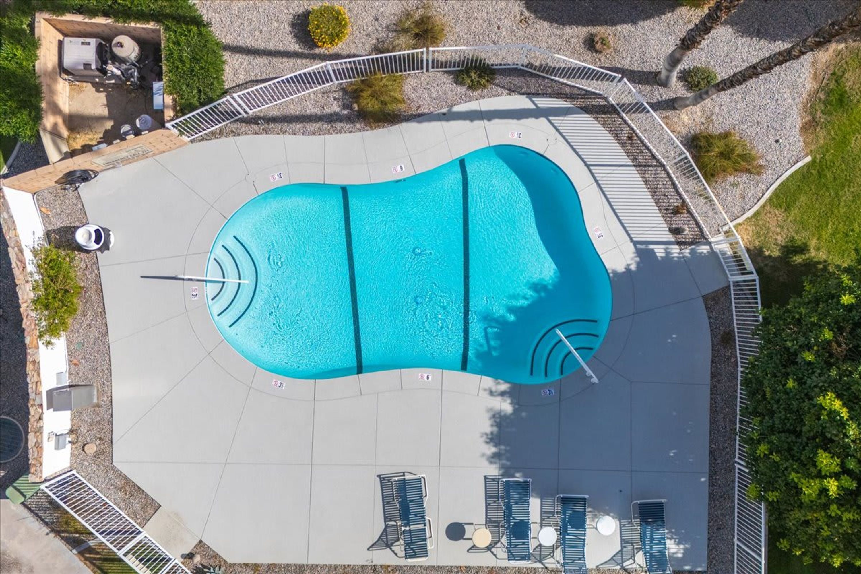 An aerial view of a swimming pool with a surrounding deck, lounge chairs, and a fenced area.
