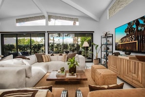Ein modernes Wohnzimmer mit einer Eckcouch, einem Couchtisch und einem Fernseher auf einer Holzkommode. Große Fenster bieten Blick auf einen Pool und Grünflächen.
