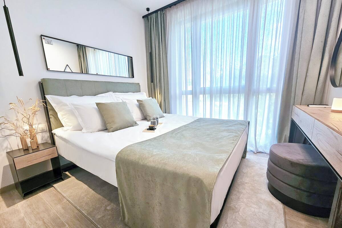 A modern hotel room with a large bed, bedside tables, a desk, and a large window with curtains.