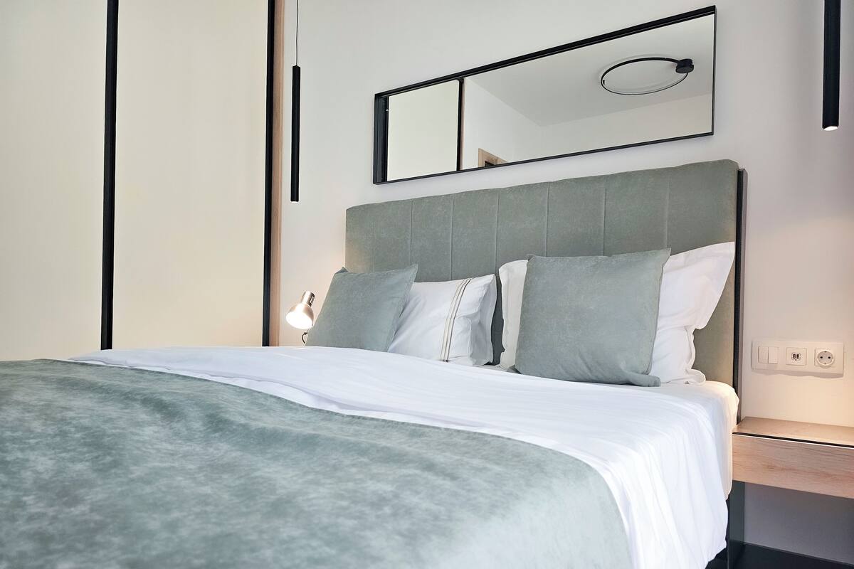 A modern hotel room with a bed, a nightstand, a lamp, and a wall-mounted mirror.