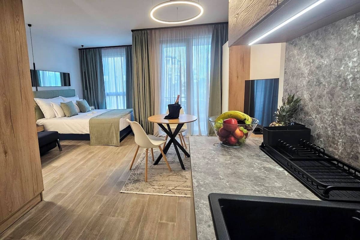 A modern hotel room with a bed, a dining table with chairs, a fruit bowl, a small kitchen area, and a TV mounted on the wall.