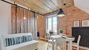 A cozy room with a white sofa, wooden ceiling, brick wall, and a dining area with a white table and chairs. - Flatbook - In the Heart of Old Town (Gdansk)