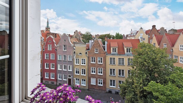 A row of colorful historic buildings with red and yellow facades, a clear blue sky, and a tree in the foreground. - Flatbook - In the Heart of Old Town (Gdansk)