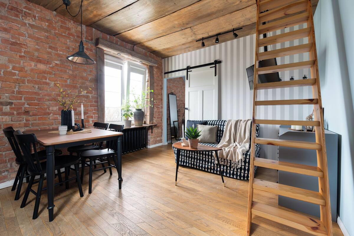 Una sala de estar acogedora con una mesa de comedor de madera, sillas negras, un sofá a cuadros blanco y negro, una escalera de madera y una pared de ladrillo.
