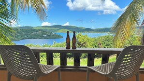 A balcony with two white chairs and two bottles of beer, overlooking a scenic view of a lake and hills.