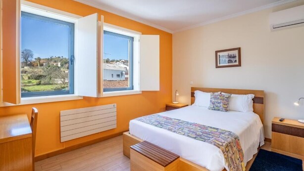Ein Schlafzimmer mit einem Bett, Nachttischen, einem Schreibtisch und einem Fenster mit geöffnetem Rollo.