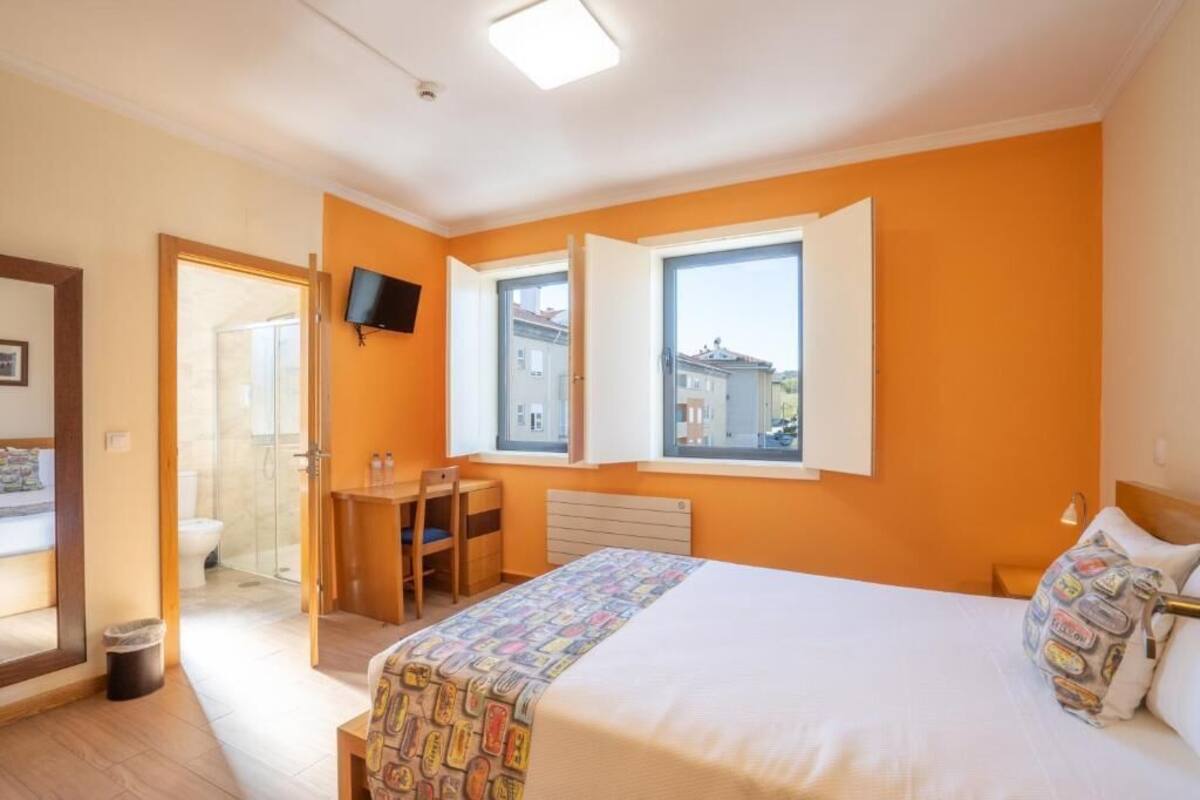 Een slaapkamer met een bed, een bureau en een stoel. Er is een raam met een open luik, een aan de muur bevestigde televisie en een deur naar een badkamer.