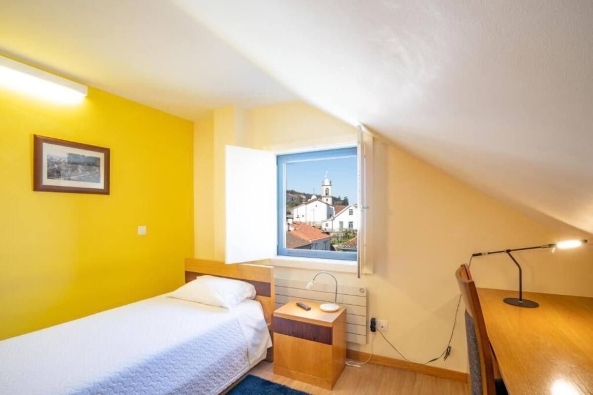 Een kleine, lichte kamer met een bed, een bureau en een raam met uitzicht op gebouwen.