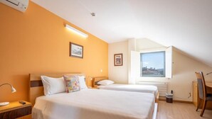 Ein Hotelzimmer mit zwei Betten, einem Schreibtisch, einem Stuhl und einem Fenster mit Blick auf die Stadt.