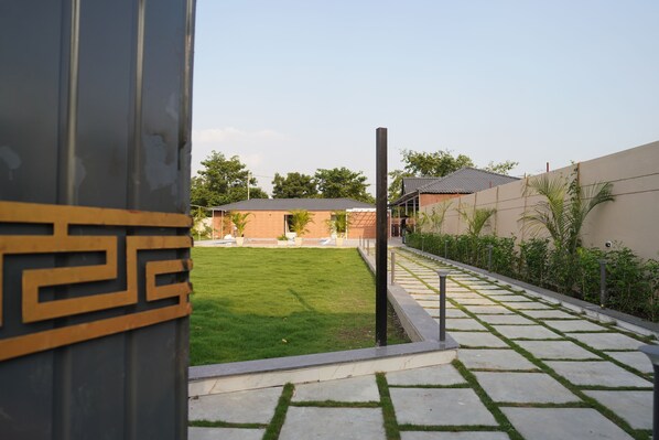A courtyard with a paved walkway, a grassy lawn, and a building with a tiled roof. - Great Escape Club (Sanwer)
