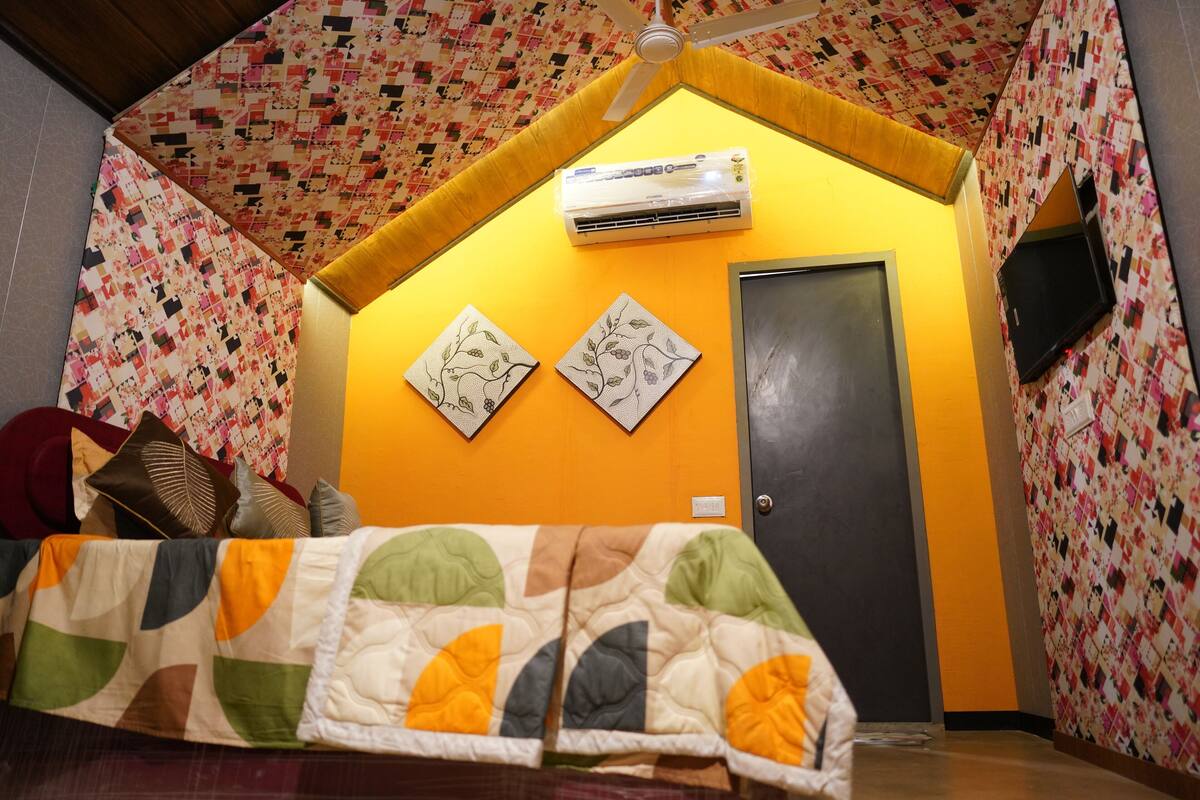 A room with a quilted bedspread, a ceiling fan, an air conditioning unit, and two framed artworks on the wall.