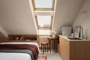 Ein kompakter, moderner Raum mit Dachfenster, einem Bett, einem kleinen Tisch mit Stuhl, einem Heizkörper und einem Waschbecken.