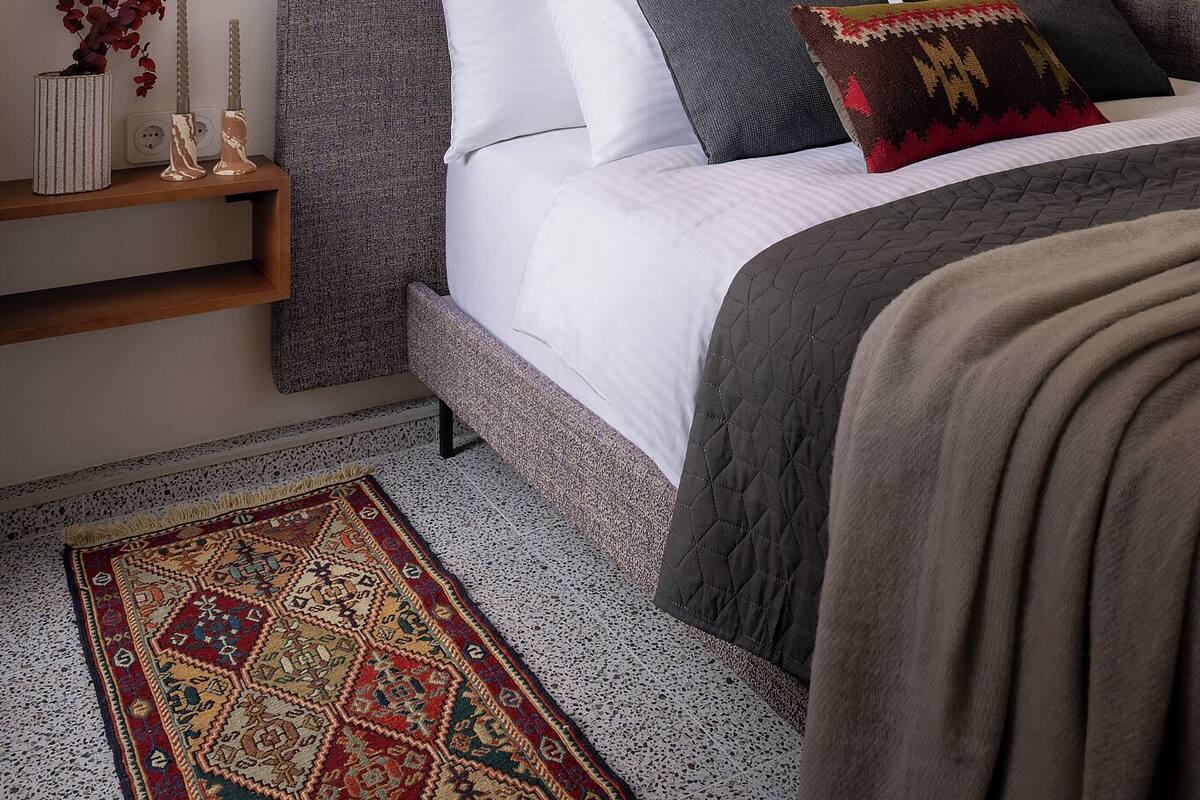A bedroom with a bed, pillows, a bedside table with a vase and candles, and a patterned rug.