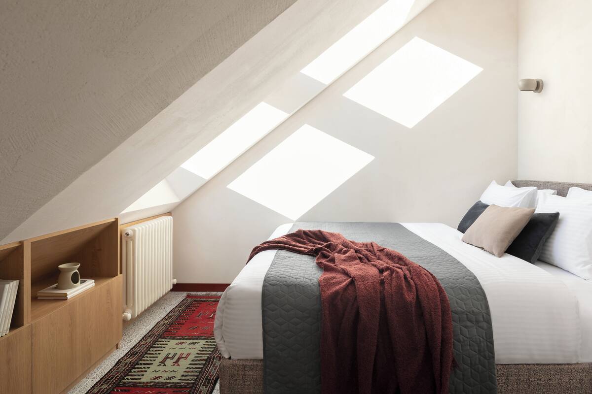 A bedroom with a sloped ceiling, skylights, a bed with pillows, a bedside table, and a radiator.