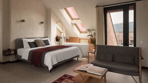 Un dormitorio moderno con cama, sofá, mesa de lectura, tragaluz y vistas a edificios.