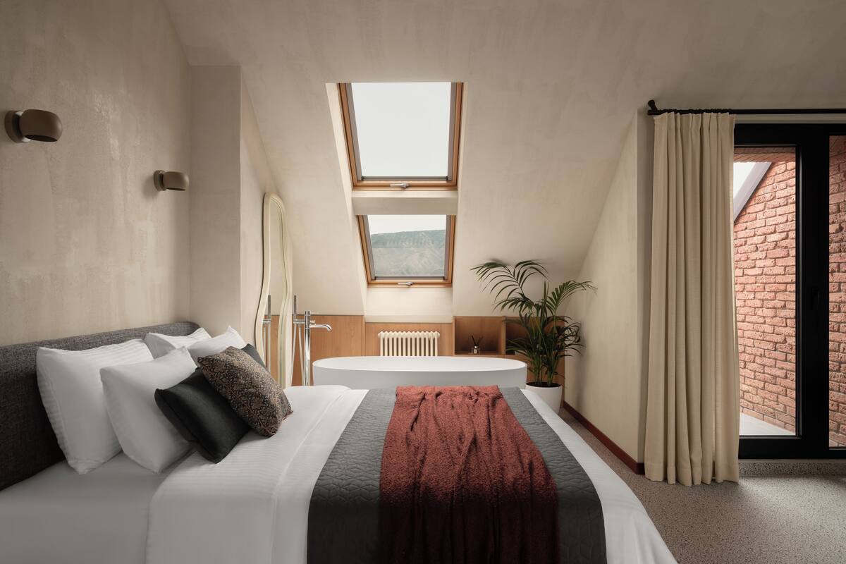 A modern bedroom with a large bed, a skylight, a bathtub, and a view of a brick wall.