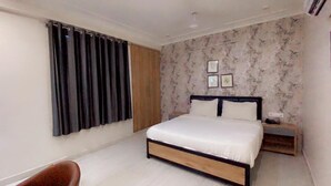 A bedroom with a bed, a nightstand, a chair, and a window with curtains. - Hotel Majesty inn (Jaipur)