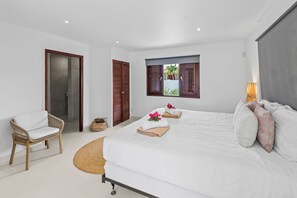 A modern bedroom with a large bed, a wicker chair, and a view of the outdoors through a window with wooden shutters.