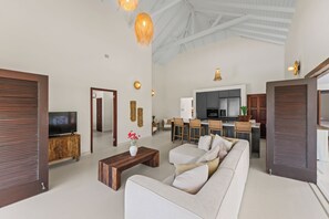 Ampla sala de estar com um sofá grande, uma mesa de centro e uma televisão. Há também uma cozinha com mesa e cadeiras.