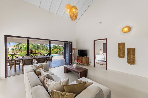 A spacious living area with a sofa, a coffee table, and a television. There is a balcony with outdoor furniture and a view of greenery.