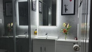 Ett modernt badrum med en duschkabin i glas, en vit tvättställskommod med en spegel ovanför och en toalett. På väggen hänger inramade tavlor och på tvättställskommoden står en vas med blommor.