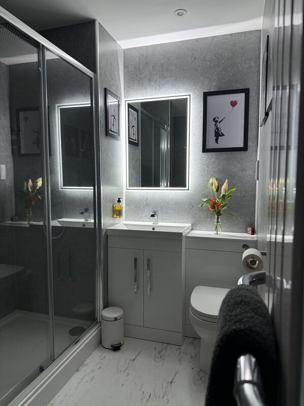 Une salle de bain moderne avec une douche à parois de verre, un lavabo blanc avec un miroir au-dessus, et des toilettes. Des tableaux encadrés ornent le mur, et un vase rempli de fleurs est posé sur le comptoir du lavabo.
