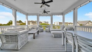 Una terraza techada al aire libre con mobiliario de mimbre blanco, ventiladores de techo y vista a edificios residenciales al fondo.