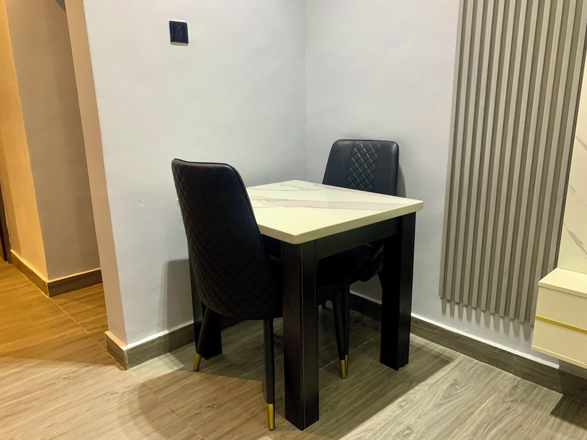 A corner room with a small table and two chairs, a window with blinds, and a wooden floor.