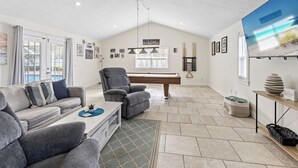 Ein großzügiges Wohnzimmer mit Blick auf einen Pool, grau bezogene Sofas, einem Billardtisch und einem großen Fernsehbildschirm.