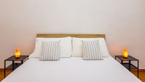 A neatly made bed with white bedding and striped pillows, flanked by two black nightstands with glowing salt lamps.