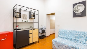 A compact kitchenette with a sink, a small fridge, and a microwave. There is a sofa with a patterned cover and a framed picture of the moon on the wall.