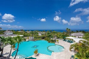 Una piscina de resort con vista al océano, rodeada de palmeras y edificios del resort.