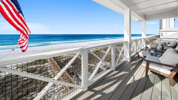 A covered deck with a view of the beach and ocean, featuring a flag, outdoor seating, and a railing. - Blessing At Pier Park 6 Bedroom Home (Panama City Beach)