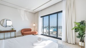 A modern hotel room with a large bed, a desk, a chair, and a balcony with a view of the water. - Sumptuous 5BR with Assist Rm in Al Dana Island, Sharm, Fujairah by Deluxe Holiday Homes (Fujairah)