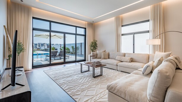 A modern living room with a large sofa, a coffee table, and a flat-screen TV. There is a large window with a view of a swimming pool and buildings. - Sumptuous 5BR with Assist Rm in Al Dana Island, Sharm, Fujairah by Deluxe Holiday Homes (Fujairah)