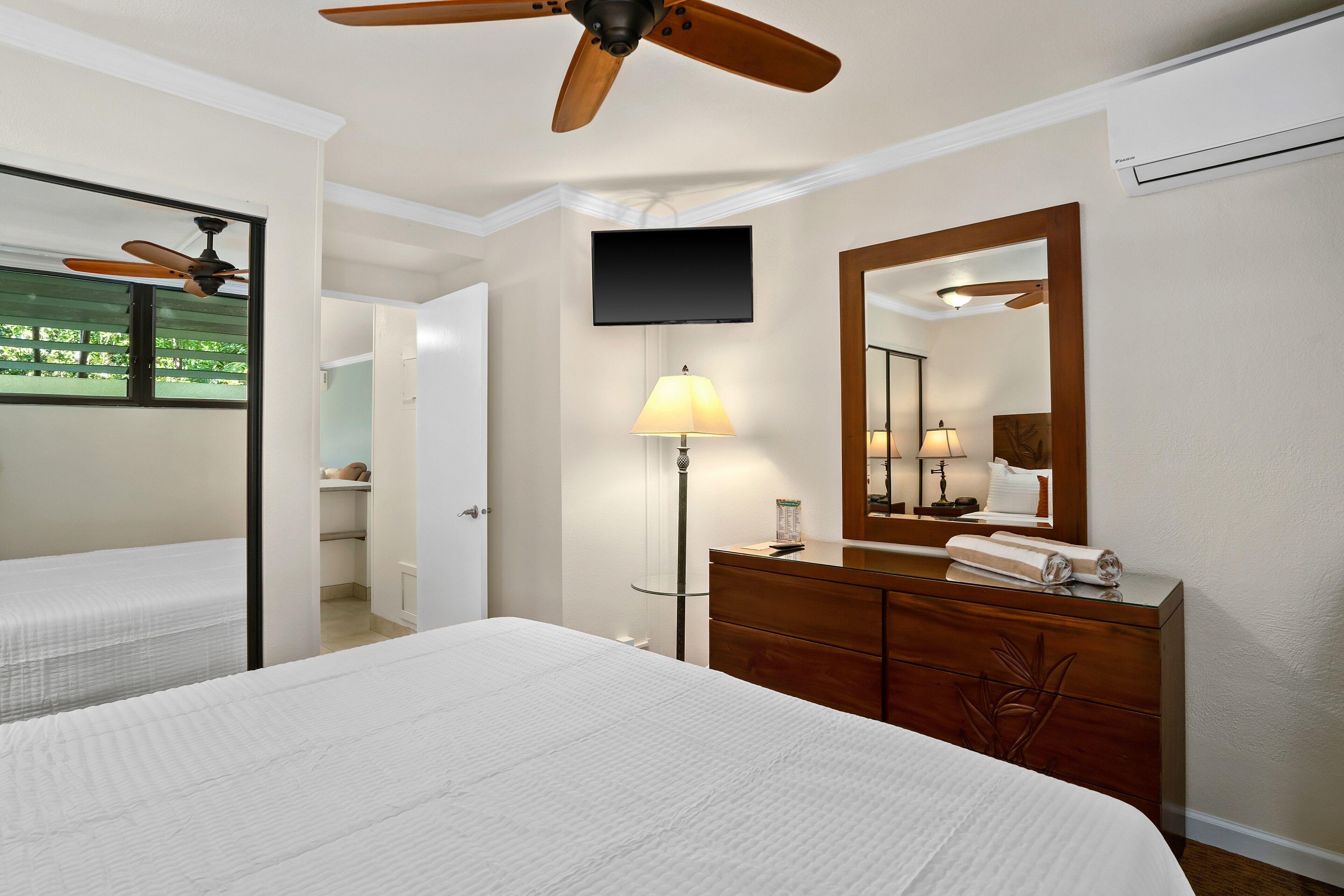A hotel room with a large bed, a wooden dresser, a mirror, a ceiling fan, a TV, and a lamp.