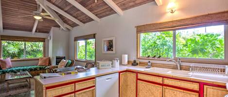 A kitchen with a wooden ceiling, a large window with a view of greenery, a built-in dishwasher, and a countertop with various appliances.