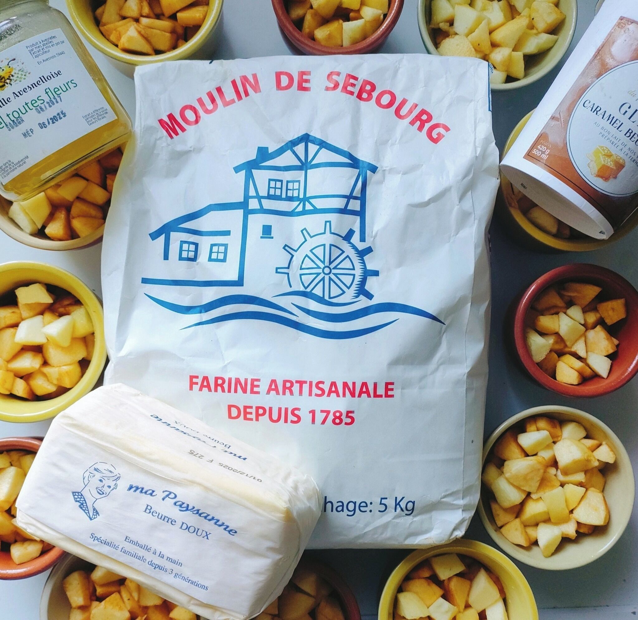 Un sac de farine artisanale de la marque « Moulin de Sebourg », orné d’un logo représentant une maison et un moulin à vent, entouré de bols remplis de pommes de terre coupées en dés.