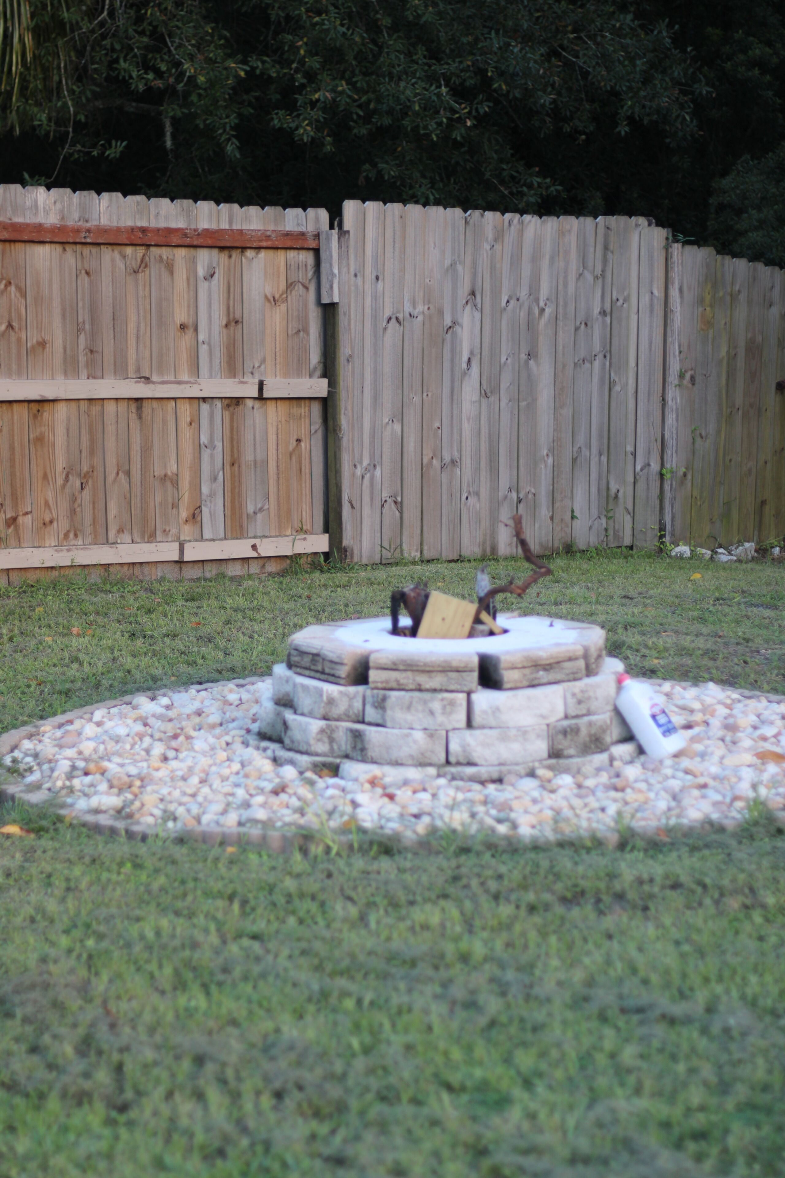 A circular stone fire pit surrounded by a gravel area, with a wooden fence in the background.